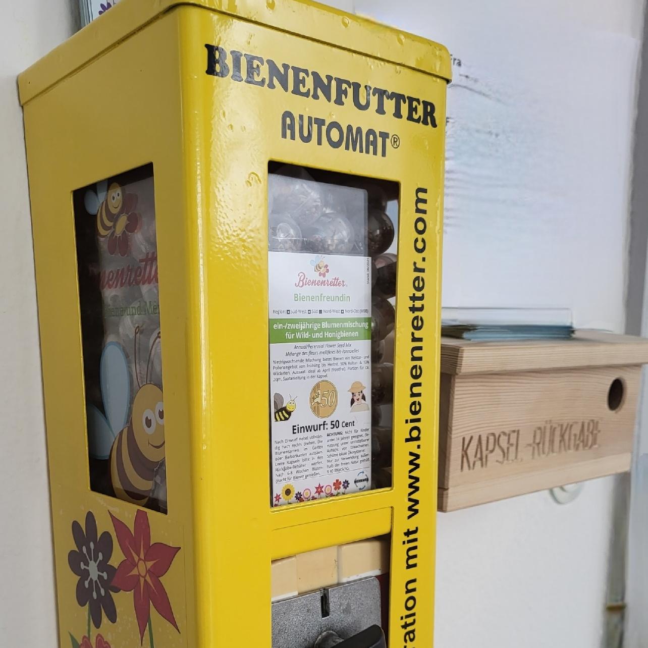 Sie sehen ein Bild unseres Bienenfutterautomaten, der aufgearbeitet wurde aus alten Kaugummiautomaten. Der Automat ist gelb angestrichen und mit Bienen und bunten Blumen bemalt.